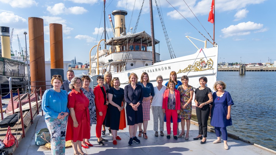 Die Minister:innen, die an der Konferenz der Gleichstellungs- und Frauenministerinnen und -minister, -senatorinnen und -senatoren der Länder teilnehmen, stehen aufgereiht vor einem Boot am Hamburger Hafen.