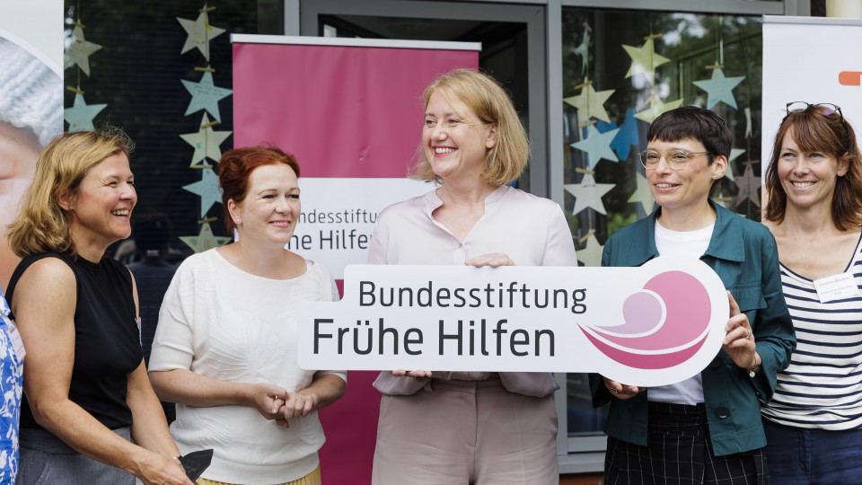 Vor Bannern der Bundesstiftung Frühe Hilfen stehen fünf Frauen, darunter Bundesfamilienministerin Paus und NRW-Familienministerin Josefine Paul, die das Logo der Bundesstiftung Frühe Hilfen in der Hand halten.