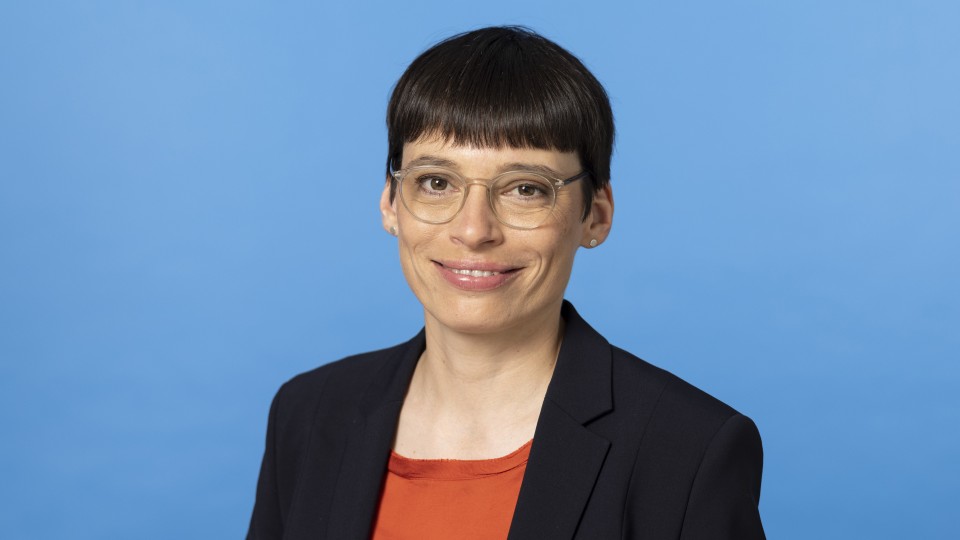 Ministerin Paul steht vor einem blauen Hintergrund. Sie lacht in die Kamera. Ein Teil des Oberkörpers ist zu sehen, der ein wenig zur Seite gewandt ist.