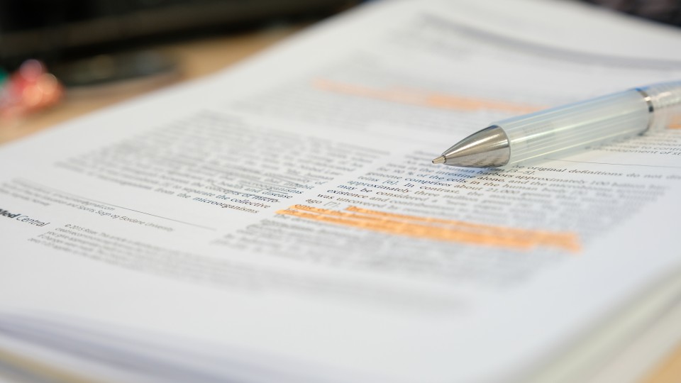 Es sind mehrere Blätter übereinander zu sehen mit Texten. Teile des Textes sind markiert. Auf den Blättern liegt ein Kugelschreiber.