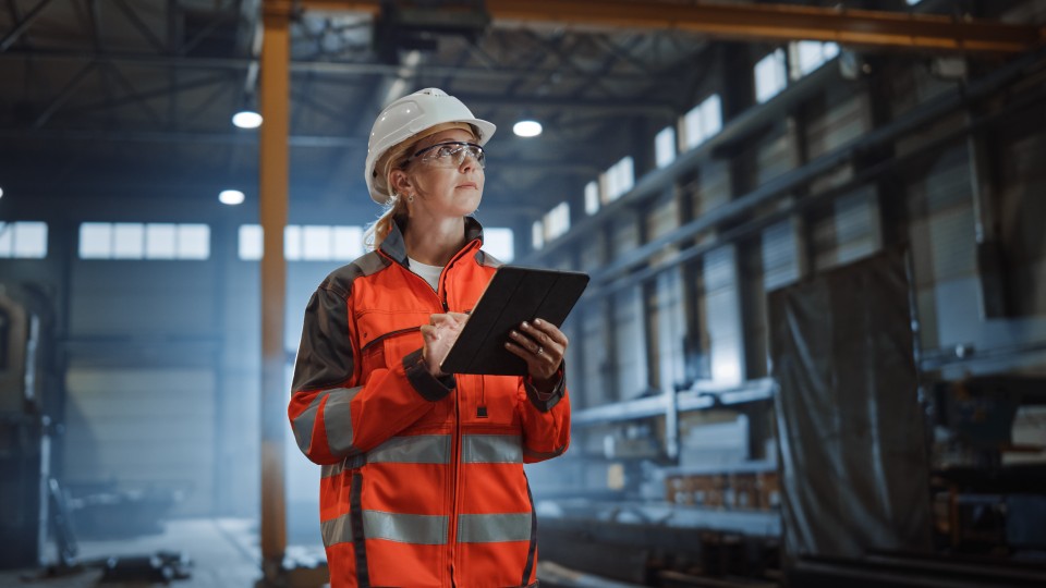 Eine Frau in orangener Warnjacke, Schutzhelm und Schutzbrille hält ein Klemmbrett in der Hand und schreibt etwas darauf. Die Frau ist in einer Industriehalle.