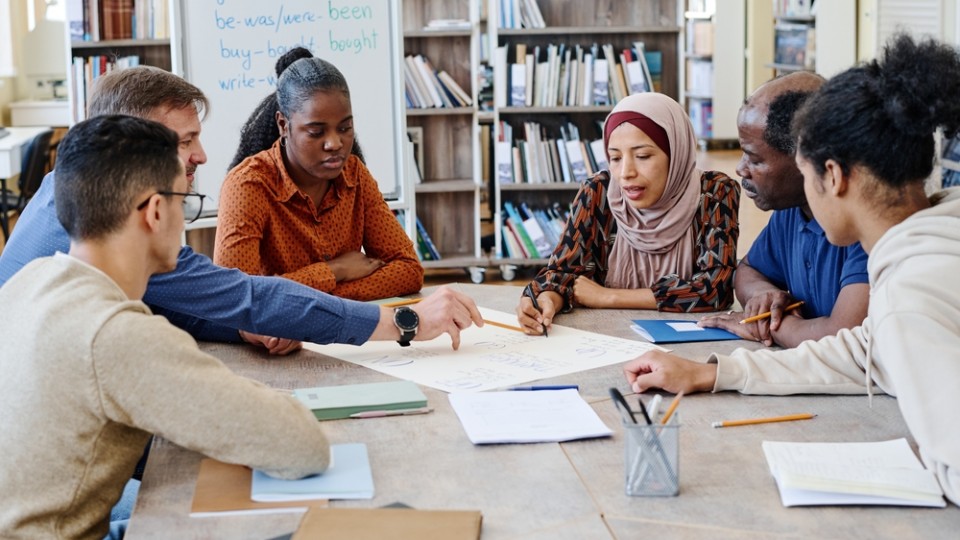 Eine Gruppe unterschiedlicher Menschen sitzen an einem runden Tisch und schauen sich gemeinsam ein großes Blatt an. Eine Person sagt etwas zu dem Blatt. Im Hintergrund stehen ein beschriebenes Flipchart und einige Regale, die mit Büchern gefüllt sind.