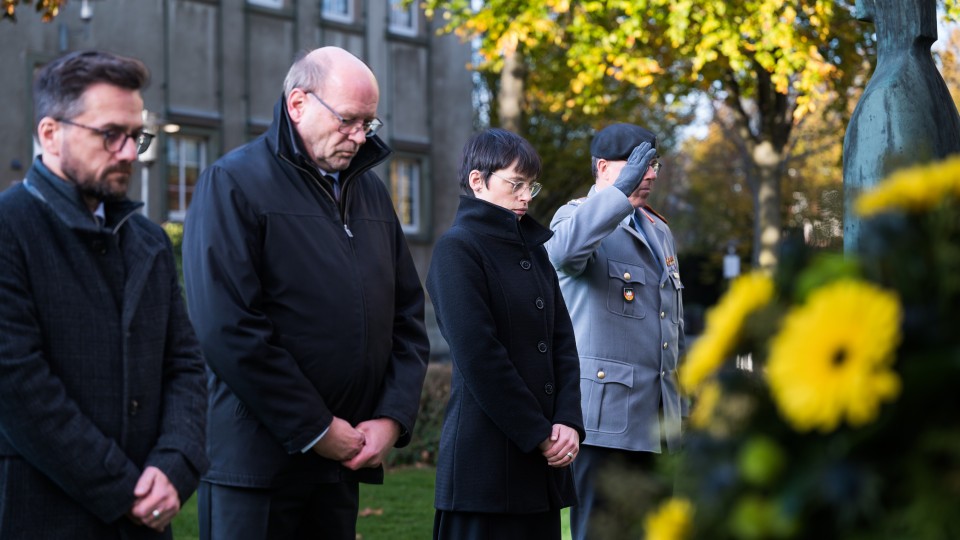 Ministerin Josefine Paul als Vertreterin der Landesregierung, Thomas Kutschaty, Staatsminister a.D., MdL, Vorsitzender des Landesverbandes NRW im Volksbund Deutsche Kriegsgräberfürsorge e.V, Rainer Schmeltzer, Landtagsvizepräsident und ein Mensch von der Bundeswehr nehmen an der gemeinsamen Gedenkveranstaltung zum Volkstrauertag teil. Sie stehen aufgereiht nebeneinander und nehmen an der stillen Kranzniederlegung teil.