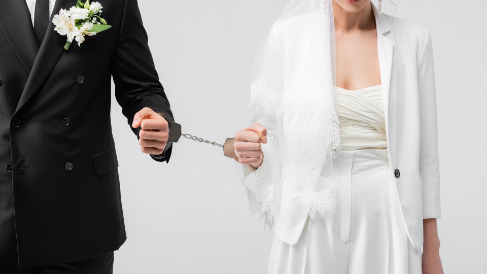 Eine Frau im Brautkleid ist mit Handschellen an einen Mann im Anzug gefesselt.