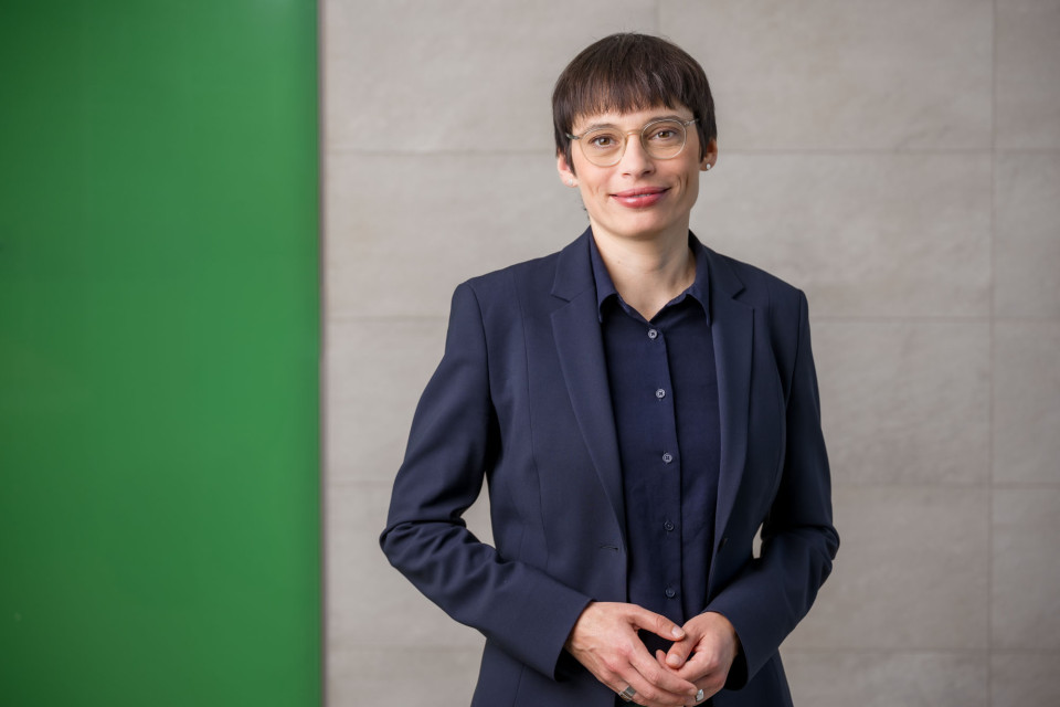 Ministerin Josefine Paul schaut lächelnd in die Kamera. Die Arme sind vor dem Körper. Die Wand im Hintergrund ist grün und beige.