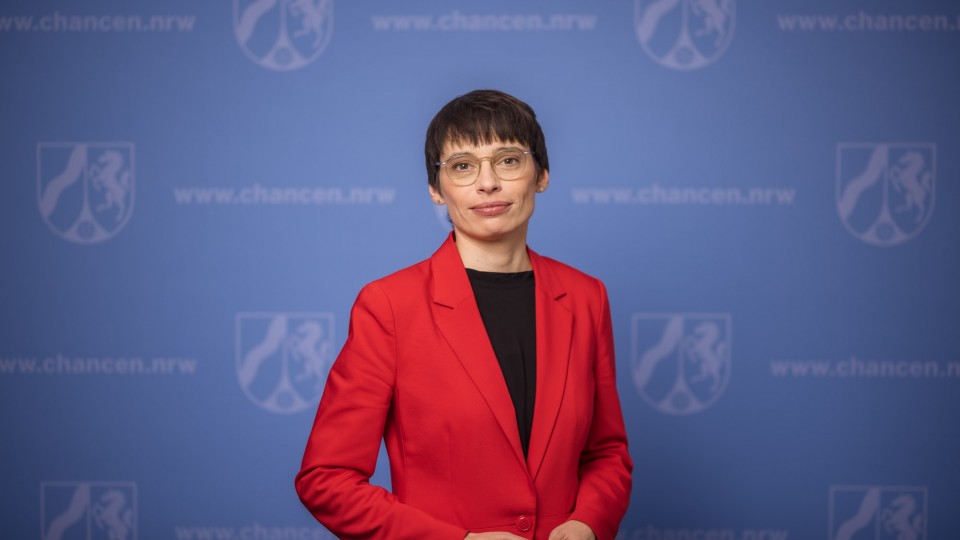 Ministerin Josefine Paul steht vor einer blauen Pressewand und schaut mit einem leichten Lächeln in die Kamera. Der Oberkörper steht leicht schräg nach rechts.