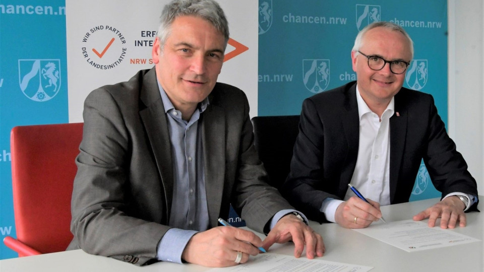 Staatssekretär Lorenz Bahr sitzt mit dem Vorstand der Caritasverband Hamm e. V., Elmar Marx, zusammen an einem Tisch. Beide unterschreiben die Partnerschaftsvereinbarung für die Landesinitiative „Erfolgsfaktor Interkulturelle Öffnung – NRW stärkt Vielfalt!“. Im Hintergrund sind die Pressewämde des Ministeriums.