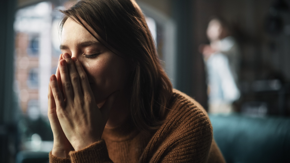 Auf dem Bild ist eine Frau zu sehen, die sich verzweifelt die Hände vor dem Gesicht verschränkt. Im Hintergrund ist unscharf ein Mann zu erkennen, der auf die Frau zeigt.