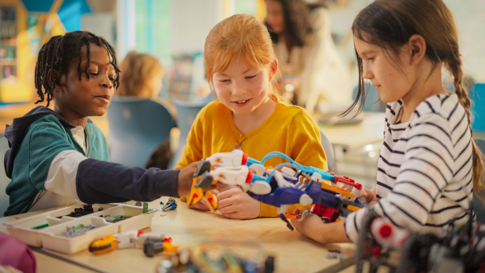 Drei Kinder bauen gemeinsam einen bunten Roboterarm in einem Klassenraum