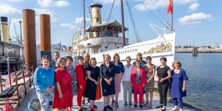 Die Minister:innen, die an der Konferenz der Gleichstellungs- und Frauenministerinnen und -minister, -senatorinnen und -senatoren der Länder teilnehmen, stehen aufgereiht vor einem Boot am Hamburger Hafen.