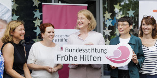 Vor Bannern der Bundesstiftung Frühe Hilfen stehen fünf Frauen, darunter Bundesfamilienministerin Paus und NRW-Familienministerin Josefine Paul, die das Logo der Bundesstiftung Frühe Hilfen in der Hand halten.