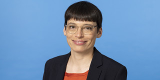 Ministerin Paul steht vor einem blauen Hintergrund. Sie lacht in die Kamera. Ein Teil des Oberkörpers ist zu sehen, der ein wenig zur Seite gewandt ist.