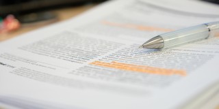 Es sind mehrere Blätter übereinander zu sehen mit Texten. Teile des Textes sind markiert. Auf den Blättern liegt ein Kugelschreiber.