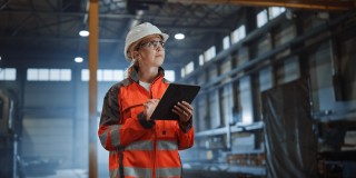 Eine Frau in orangener Warnjacke, Schutzhelm und Schutzbrille hält ein Klemmbrett in der Hand und schreibt etwas darauf. Die Frau ist in einer Industriehalle.