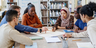 Eine Gruppe unterschiedlicher Menschen sitzen an einem runden Tisch und schauen sich gemeinsam ein großes Blatt an. Eine Person sagt etwas zu dem Blatt. Im Hintergrund stehen ein beschriebenes Flipchart und einige Regale, die mit Büchern gefüllt sind.