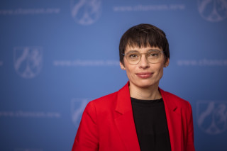 Frau Paul steht vor einer blauen Pressewand. Sie schaut in die Kamera mit einem leichten Lächeln. Der Oberkörper ist nur angeschnitten.