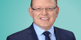 Ein Mann im Anzug lächelt vor einem grünen Hintergrund.