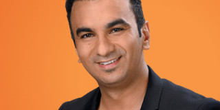 Ein Mann lächelt vor einem orangefarbenen Hintergrund. Formelle Kleidung, freundlicher Ausdruck.