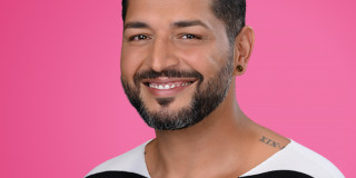 Lächelnder Mann vor rosa Hintergrund, trägt gestreiftes Oberteil, hat Bart und Ohrschmuck.