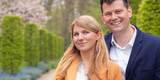 Zwei lächelnde Personen in einem gepflegten Garten mit grünen Hecken.