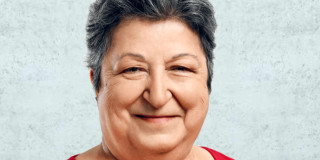 Ältere Frau lächelt freundlich in die Kamera, trägt ein rotes Oberteil.
