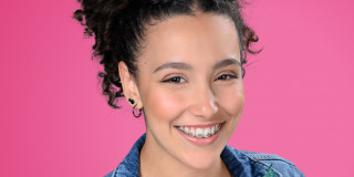 Lächelnde Person mit Locken und Jeansjacke voller bunter Anstecker vor pinkem Hintergrund.