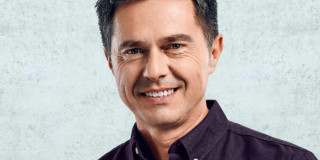 Ein Mann lächelt freundlich, trägt ein dunkles Hemd vor hellem Hintergrund.