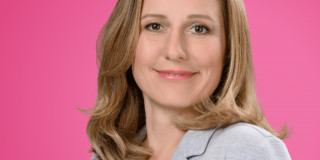 Frau mit braunen Haaren lächelt vor pinkem Hintergrund, trägt hellen Blazer.