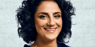 Eine lächelnde Person mit lockigem Haar trägt eine blaue Jacke vor hellem Hintergrund.