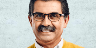 Ein Mann mit Brille lächelt, trägt gelben Pullover und weißen Hemdkragen.