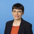 Ministerin Paul steht vor einem blauen Hintergrund. Sie lacht in die Kamera. Ein Teil des Oberkörpers ist zu sehen, der ein wenig zur Seite gewandt ist.