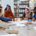 Eine Gruppe unterschiedlicher Menschen sitzen an einem runden Tisch und schauen sich gemeinsam ein großes Blatt an. Eine Person sagt etwas zu dem Blatt. Im Hintergrund stehen ein beschriebenes Flipchart und einige Regale, die mit Büchern gefüllt sind.