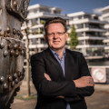Der Poloniabeauftragte Dr. Andreas Hollstein posiert für ein Foto. Er steht mit voreinander verschränkten Armen und leicht zur Seite gedrehtem Körper vor Häusern, die im Hintergrund zu sehen sind.
